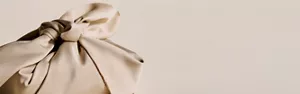 Nahaufnahme der Hand einer Person, die einen Knoten an einem beigen Furoshiki-Tuch bindet, vor einem beigen Hintergrund